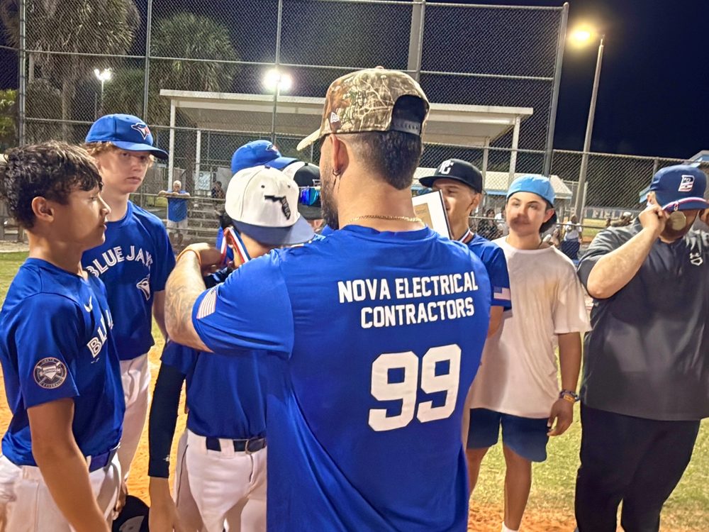 A coach in a Nova Electrical Contractors jersey talking with young baseball players